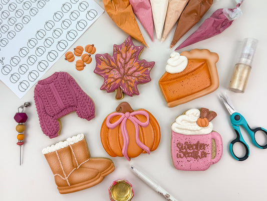 Decorative cookies shaped like a sweater, pumpkin, leaf, pie, and mug with autumn-themed decorations on a white surface.